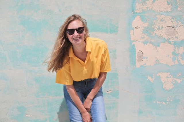 Žena v žltom tričku so slnečnými okuliarmi stojí pri stene.jpg